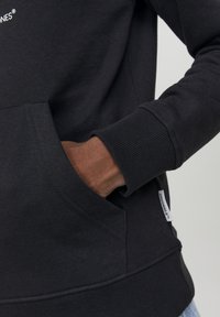 Sudadera negra con puños y dobladillo acanalados, que cuenta con un bolsillo en la parte frontal y una etiqueta de tela en el lateral. Material suave y texturizado.