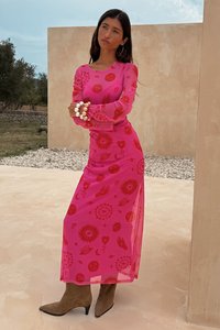Vestido maxi rosa com padrões gráficos vermelhos, mangas longas e transparentes, uma fenda lateral, combinado com botas de cano curto castanhas e uma pulseira de pérolas.