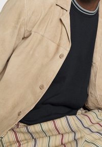 Béžová semišová bunda s button zapínáním, nošená přes tmavý svetr. Pruhované kalhoty mají více barev a jemnou texturu.