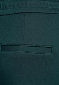 Tmavě zelená, pružná tkanina s hladkým povrchem; obsahuje zadní kapsu s obdélníkovým klopovým víkem a viditelným prošíváním podél okraje.