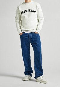 Felpa bianca con testo nero "PEPE JEANS", abbinata a jeans blu larghi e sneakers bianche, creando un outfit casual.