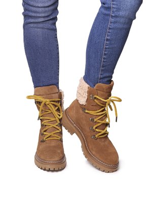 Bottes chelsea en suède marron avec des lacets jaunes, doublure en fausse fourrure et semelles en caoutchouc robustes. Conçues pour le confort et l'adhérence.