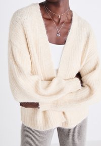 Cream-colored oversized cardigan with a soft, fuzzy texture, deep V-neck, and ribbed detailing. Paired with a gray ribbed pant.