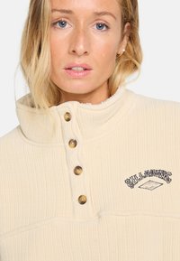 Crème kleurige pullover met een hoge kraag, geribbelde textuur en vier bruine knopen. Heeft een geborduurd logo detail op de borst.