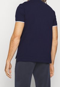 Mörkblå pikétröja med korta ärmar, vita detaljer på ärmarna och kragen, samt sidoförslitningar i nederkanten för komfort och passform.