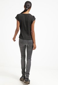 Schwarze Kurzarmbluse mit gerafftem Schulterdetail, kombiniert mit grauen Skinny-Jeans und schwarzen Schuhen, Ansicht von hinten.