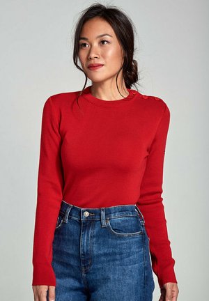 Rode ribgebreide trui met lange mouwen, een ronde halslijn en knoopaccenten op de schouder, gecombineerd met blauw high-waisted jeans.