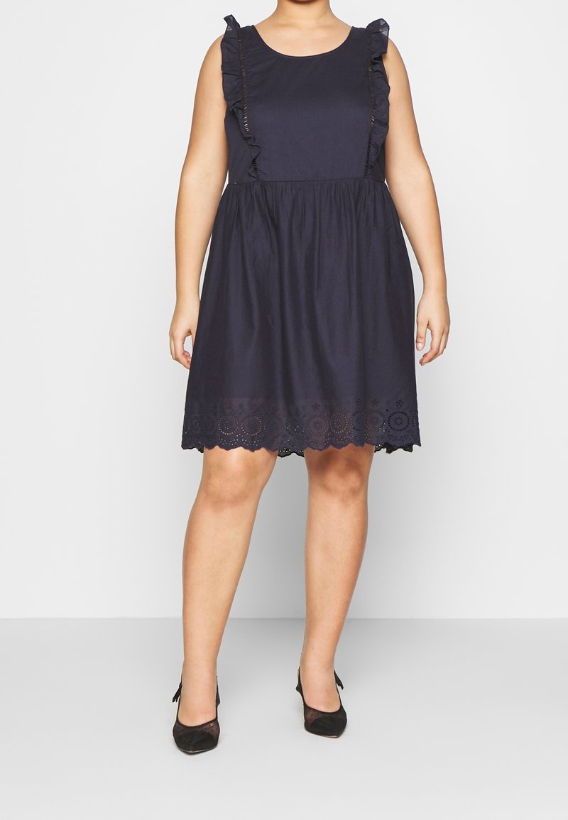 Robe noire sans manches longueur genou avec bretelles à volants et ourlet festonné portée avec des talons noirs à bout pointu sur une personne debout.