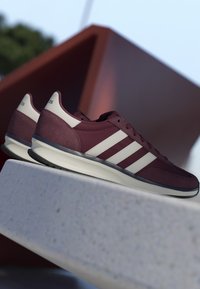 Chaussures de sport bordeaux avec des bandes blanches et des semelles Cloudfoam reposant sur un rebord en béton gris devant un arrière-plan extérieur flou.