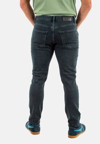 Jeans in denim blu scuro con una vestibilità slim, dotati di due tasche posteriori e un tessuto testurizzato. Indossati con calzature blu e una camicia verde.