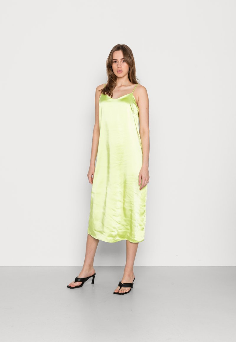 Vestido slip de cetim verde limão com alças finas, comprimento até o joelho, textura suave e ligeiro brilho, usado com sandálias de salto preto com tiras.