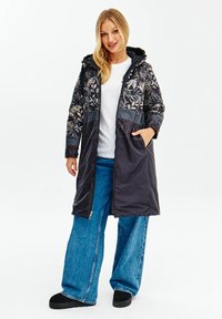 Dlouhý černý zateplený kabát s květinovým vzorem nahoře, na zip, s kapucí a kapsami. Nosený přes bílou košili a volné modré džíny.