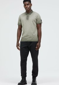 Grüne Polohemd mit kurzen Ärmeln, strukturiertem Stoff, zwei Knöpfen und einem dunklen Kragen. Kombiniert mit schwarzen Hosen und schwarzen Schnürstiefeln.