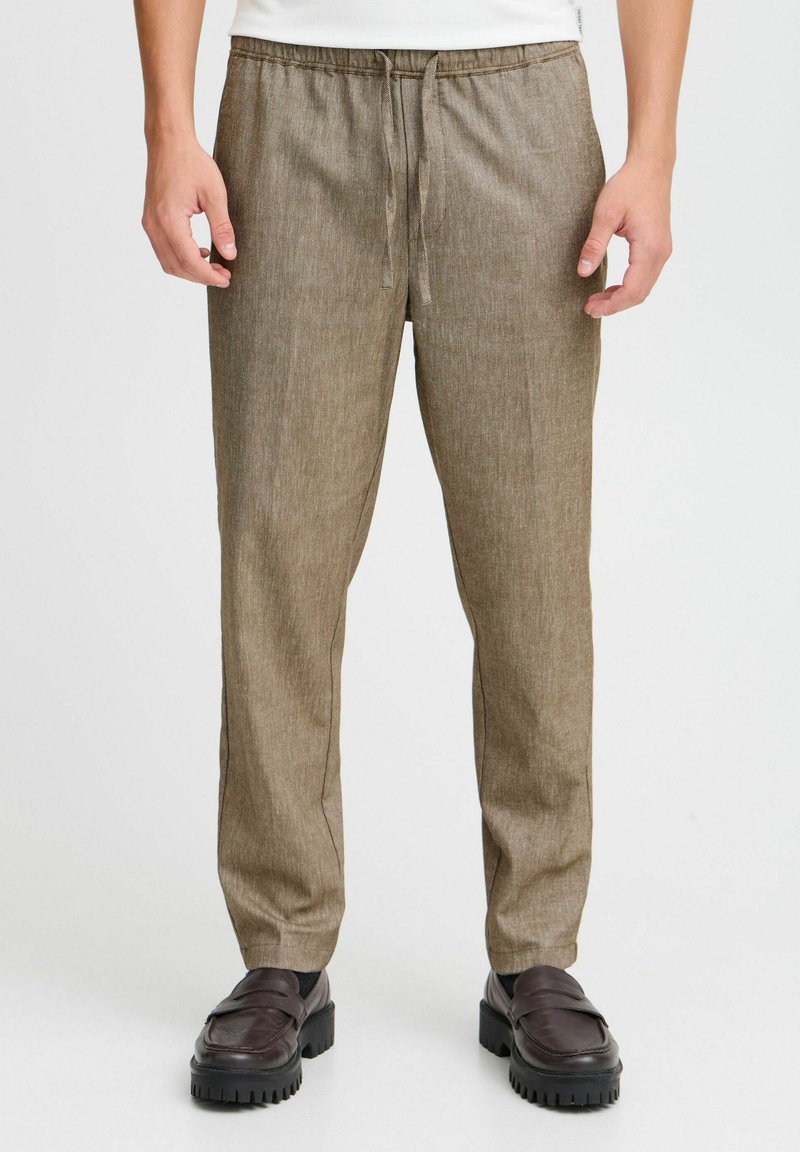Man wearing light brown drawstring pants and dark brown chunky loafers, standing against a white background.