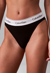 Frau trägt schwarze Calvin Klein Unterwäsche mit weißem Bund, auf dem der Markenname in schwarzen Buchstaben zu sehen ist.