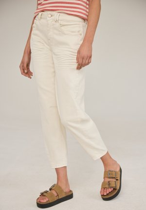 Person iført cremefarvede bukser med lige ben, striped rød og hvid skjorte og brune sandaler med doble spænder, stående mod en ensfarvet baggrund.