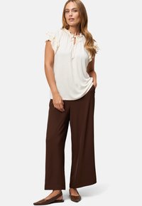 Lichtbeige top met ruches mouwen en een keyhole halslijn, gecombineerd met bruine wijde broek en platte bruine schoenen. Glad stofoppervlak.