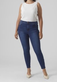 Weißes, ärmelloses Top mit Rippenmuster, kombiniert mit dunkelblauen Skinny-Jeans. Die Jeans haben einen Knopfverschluss und eine klassische Denim-Textur.