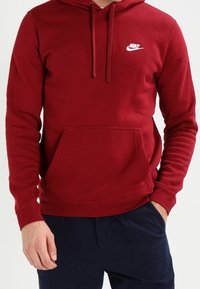 Czerwony bawełniany hoodie z kapturem ze sznurkiem, kieszenią kangurką i białym logo Nike na piersi. Prążkowane mankiety i dół.