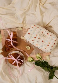 Pojemnik z pikowanej tkaniny w pastelowe wzory jedzenia w kolorze kremowym, z różowymi akcentami wstążki, dwa rogaliki na brązowym papierze i białe róże.