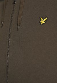Felpa con zip color marrone in tessuto a coste, con un logo di un uccello ricamato giallo sul petto e lacci intrecciati.