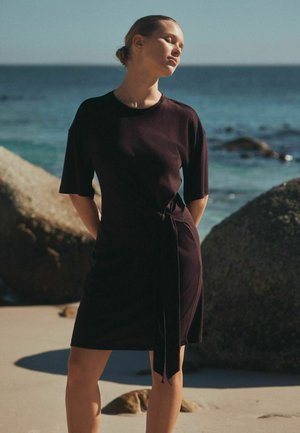 Egy nő áll homokos tengerparton közel a sziklákhoz és az óceánhoz, fekete rövid ujjú ruhát visel, oldalán megkötve, csukott szemmel, a napfény felé fordulva.