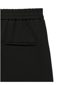 Détail d'un pantalon en tissu noir avec une ceinture élastique et une poche arrière fendue horizontalement sans fermetures visibles.