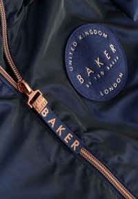 Mørkeblå stof med en glat tekstur; har et cirkulært logopatch og en lynlås med en roseguld trækflig indgraveret med "TED BAKER."