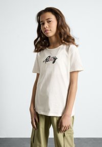 T-shirt couleur crème en coton, avec des manches courtes et un col rond, orné de texte brodé dans des teintes foncées et roses à l'avant.