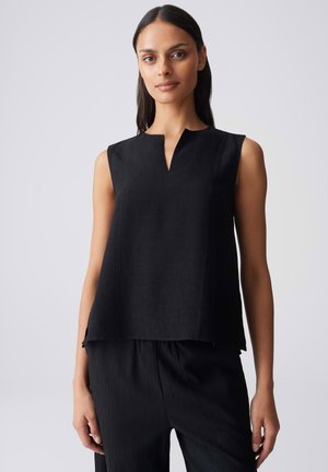 Femme aux longs cheveux foncés portant un haut noir sans manches avec un décolleté en V et un pantalon noir assorti, debout devant un fond gris.