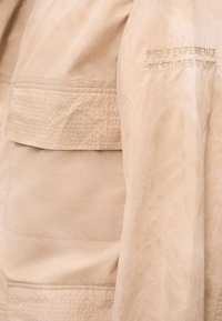 Μπεζ υφασμάτινες πτυχές με ορατή ραφή και κεντημένο κείμενο που αναγράφει «AIGLE EXPERIENCE BY ETUDES STUDIO» στη δεξιά πλευρά.