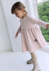 Bledoružové bavlnené šaty s dlhými rukávmi, nariasenou sukňou a kvetinovou výšivkou. Obsahujú hravú ilustráciu na leme. Kombinované s béžovými členkovými čižmami.