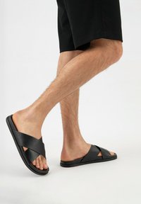 Sandales noires à glissière avec des larges brides croisées, fabriquées en matériau synthétique lisse, dotées d'une semelle plate et d'un design ouvert au niveau des orteils.