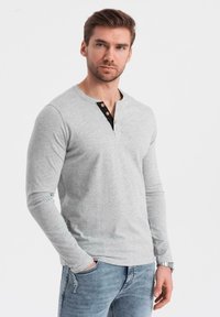 Man med kort brunt hår som bär en ljusgrå långärmad henley-tröja med svart knäppning och blå jeans, står med en hand i fickan.