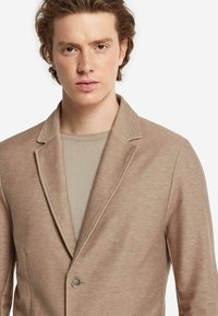 Beige kavaj med texturerad tyg, med slagkrage och enkel knappknäppning, lager på lager över en ljusbeige skjorta.
