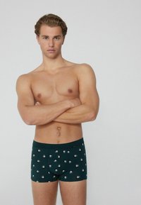 Oben ohne Mann mit verschränkten Armen, der dunkle Boxershorts trägt, die mit kleinen 8-Ball-Mustern bedruckt sind, vor einem schlichten hellen Hintergrund stehend.