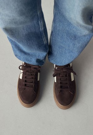 Nohy v hnědých semišových teniskách s bílými pruhy a hnědými tkaničkami, doplněné světle modrými džínami na šedé podlaze.