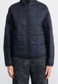 Personne portant une veste matelassée noire zippée à manches longues et un pantalon foncé à motifs, debout devant un fond clair uni.