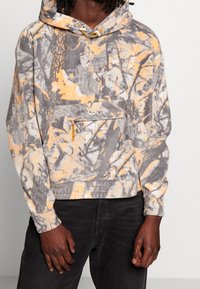Fleece hættejakke i grå, orange og hvid camouflage mønster. Har en frontlomme, justerbar snørehætte og ribbede manchetter.