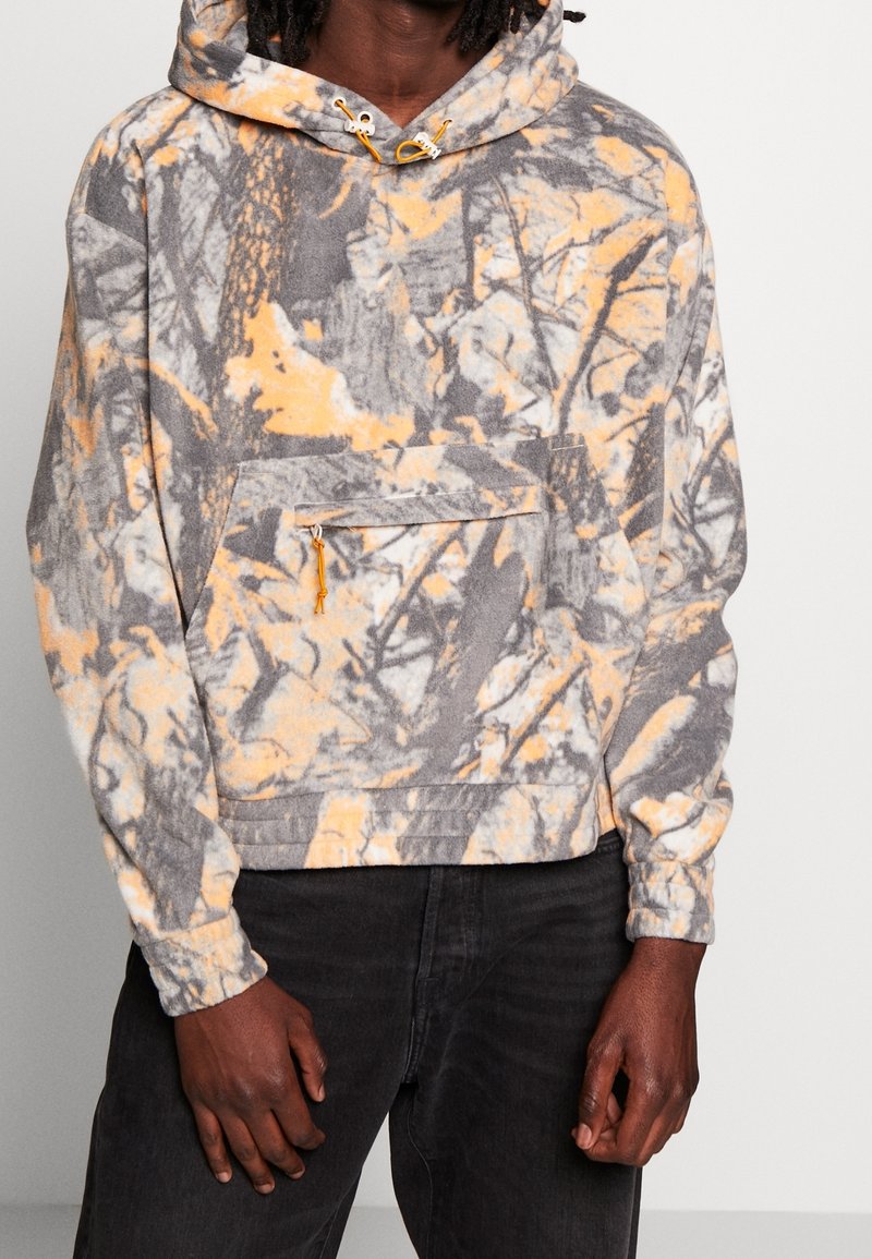 Fleece hættejakke i grå, orange og hvid camouflage mønster. Har en frontlomme, justerbar snørehætte og ribbede manchetter.