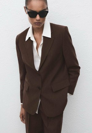 Brown tailored suit jacket with notch lapels, two front pockets, single button closure, worn over a white pinstripe shirt. Matte black sunglasses.