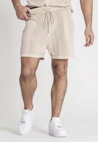 Beige gebreide shorts met opengewerkte patronen, elastische tailleband met trekkoord, zijzakken en een relaxte pasvorm, gecombineerd met witte sneakers.