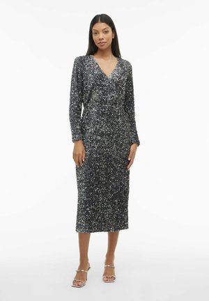 Abito nero di paillettes con maniche lunghe e scollo a V, caratterizzato da un taglio a metà lunghezza e una superficie texturizzata. Indossato con sandali argento con cinturini.