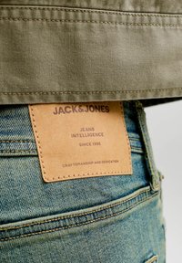 Nahast plaastrit sinistel teksapükstel, millel on kiri "JACK & JONES JEANS INTELLIGENCE ALATES 1990. AASTAST, OSAKUNSTI JA PÜHENDUNUD" rohelise kangariide all.