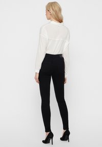 Mujer rubia con una blusa blanca de manga larga, jeans ajustados negros y zapatos de tacón altos negros, de espaldas contra un fondo liso.