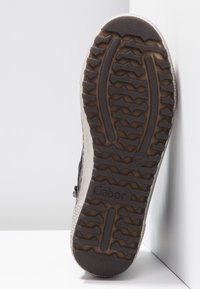 Semelle d'une chaussure Gabor avec des motifs de bande de roulement profonds en caoutchouc brun foncé, présentée à la verticale sur des surfaces blanches.
