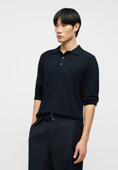 Άνδρας με μαύρα μαλλιά που φοράει μαύρο πόλο με μακριά μανίκια και μαύρο παντελόνι με λεπτές ρίγες, με τα χέρια στις τσέπες, σε απλό φωτεινό φόντο.