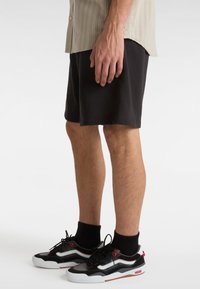 Shorts neri con una trama solida, abbinati a scarpe Vans nere e grigie con accenti rossi e suole bianche. Camicia a righe parzialmente visibile.