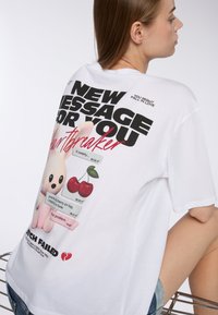 Biała, oversize'owa koszulka z kolorową grafiką z tyłu, przedstawiającą postać z kreskówki, czereśnie oraz frazy tekstowe w pogrubionej czcionce.