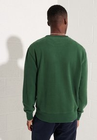 Muž s krátkými vlasy, oblečený v tmavě zelené mikině s dlouhým rukávem a tmavých kalhotách, stojí zády k světle cihlové zdi.